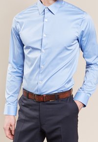 Ljusblå långärmad skjorta med krage, knappar fram och en slät textur, ihop med mörka byxor och ett brunt bälte.
