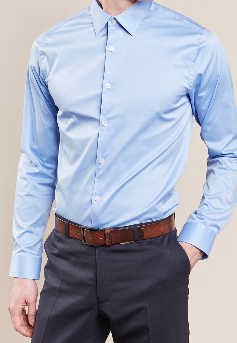 Ljusblå långärmad skjorta med krage, knappar fram och en slät textur, ihop med mörka byxor och ett brunt bälte.
