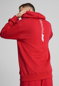 Piros kapucnis pulóver laza szabással, elől kenguruzsebbe építve, fehér "Formula 1" logóval a bal oldalon. Sima textúra.