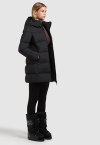 Zwarte pufferjas met een hoge kraag, voorritssluiting en horizontale gestikte textuur. Gecombineerd met zwarte leggings en winterlaarzen.