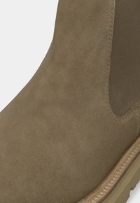 Brune ruskindsankelstøvle med en glat overflade, elastiske sidedele og en tan gummisål. Har syede detaljer omkring bunden.