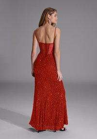 Abito da sera rosso in paillettes con corpino aderente, spalline sottili e gonna fluente. Presenta una schiena bassa e una superficie strutturata e scintillante.