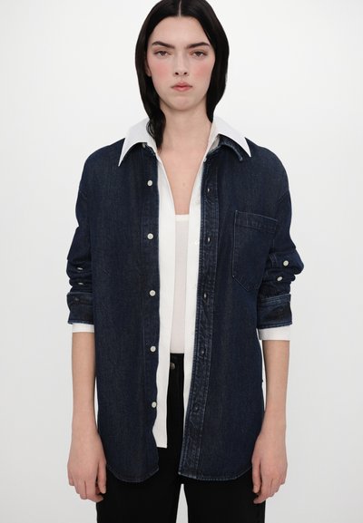 Chemise en denim foncé avec un col blanc et une fermeture à boutons. Caractéristiques : manches retroussées et une poche poitrine. Design classique.
