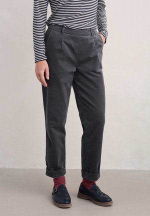 Person trägt dunkelgraue, hochgekrempelte Hose, schwarze Schnallenschuhe, burgunderfarbene Socken und ein schwarz-weiß gestreiftes Langarmshirt, steht auf einem grauen Boden.