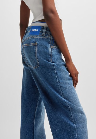 Jeans en denim bleu à jambes larges avec une taille haute, arborant une étiquette logo "HUGO" bleue à l'arrière et des détails de couture minimalistes sur les poches.