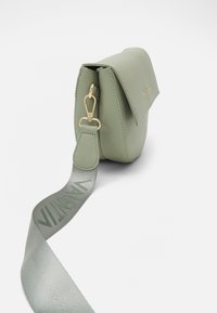 Zelená kabelka z umělé kůže přes rameno s klopovým uzávěrem, zlatými detaily a texturovaným popruhem se logem. Kompaktna obdélníková forma.