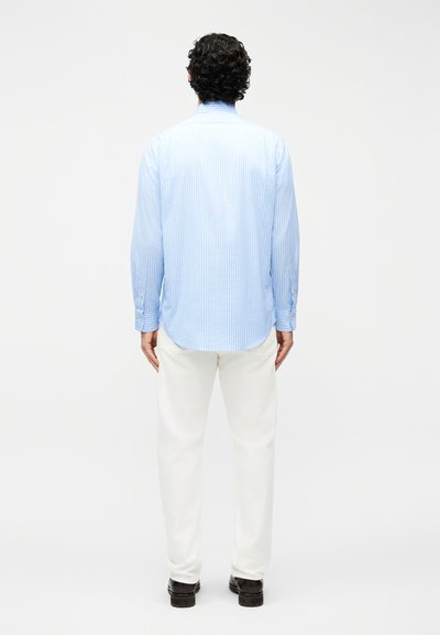 Άνδρας με σγουρά σκουρόχρωμα μαλλιά, φοράει ένα ανοιχτό μπλε ριγέ πουκάμισο με μακριά μανίκια, λευκό παντελόνι και μαύρα παπούτσια, στέκεται γυρισμένος προς τα πίσω.