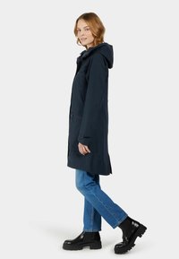 Námořnický modrý nepromokavý parka s kapucí, předními kapsami a zipy, spárovaný se světle modrými džíny a černými robustními botami.