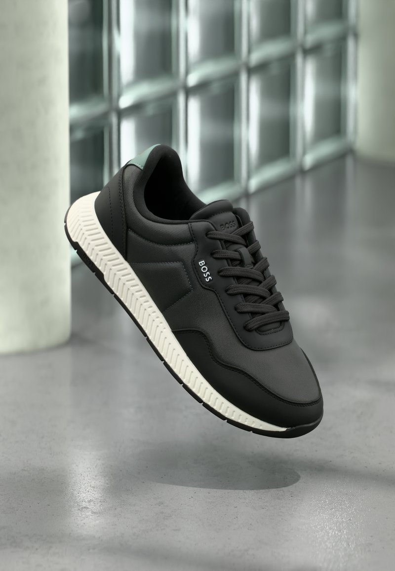 Μαύρο και γκρι αθλητικό παπούτσι με λευκή σόλα και λογότυπο "BOSS" στο πλάι και στη γλ ώσσα, αιωρούμενο πάνω από μια αντανακλαστική γκρι επιφάνεια κοντά σε τοίχο από γυάλινους κύβους.