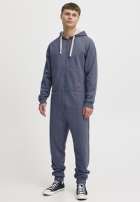Navy blue hooded onesie with a front zipper, kangaroo pocket, and beige drawstrings; features ribbed cuffs and a small logo patch.