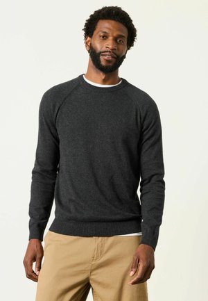Mand med skæg iført en mørkegrå langærmet sweater og beige bukser står mod en ensfarvet lys baggrund.