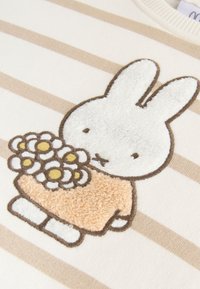 Lapin blanc aux yeux fermés tenant un bouquet de fleurs blanches et jaunes sur un tissu rayé beige et blanc.