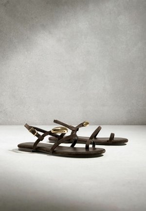 Sandale plate maro cu bretele subţiri şi ornamente ovale aurii pe bretelele de la gleznă, expuse pe o suprafaţă neutră pe un fundal gri.