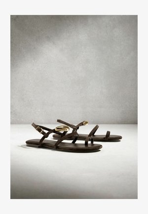 Pruunid lamedad sandaalid õhukeste rihmade ja kuldsete ovaalsete kaunistustega pahkluu rihmadel, eksponeeritud neutraalsel pinnal halli tausta ees.