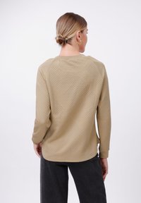 Maglietta beige a manica lunga con un motivo geometrico testurizzato, colletto rotondo e spalle cadenti, abbinata a pantaloni scuri.