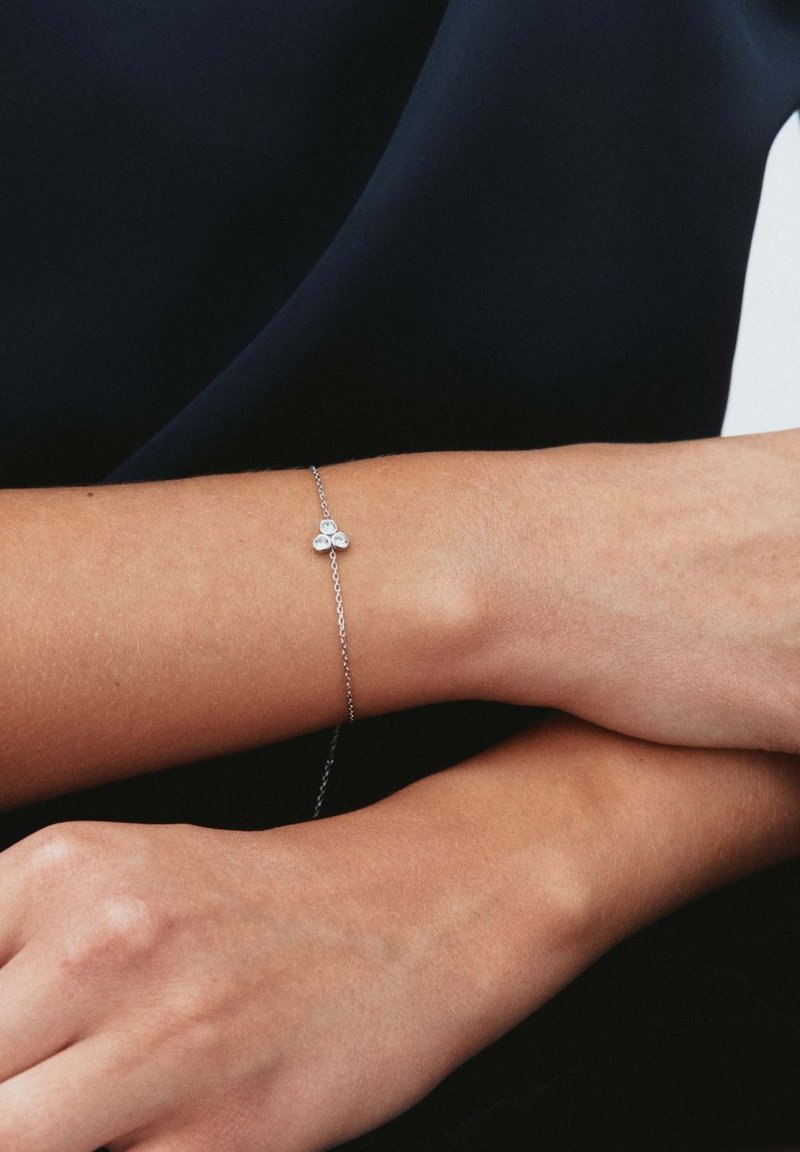 Bracciale d'argento con una delicata catena decorata da un trio di gemme circolari, posato su un polso ambrato su uno sfondo scuro.
