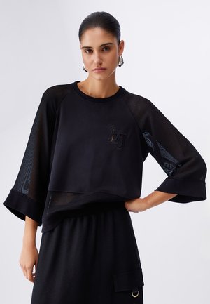 Donna con capelli lisciati all'indietro indossa una maglia nera oversize con maniche trasparenti e una gonna nera con dettaglio tasca, in piedi davanti a uno sfondo semplice.