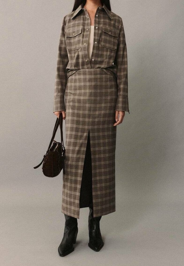 Robe chemise à carreaux marron avec une fente à l'avant, dotée de deux poches poitrine. Associée à des bottes noires montantes jusqu'au genou et un petit sac à main marron.