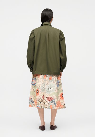 Γυναίκα όρθια πλάτη φοράει ένα μακρυμάνικο μπουφάν ελιάς και μια φούστα μέχρι το γόνατο με πολύχρωμο λουλουδάτο μοτίβο.