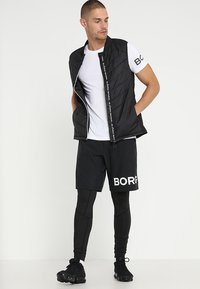 Čierna vycpaná vesta so zipsom, bielym brandingom, nosená cez biele tričko, v kombinácii s čiernymi šortkami a priliehavými legínami. Čierne topánky dokončujú vzhľad.