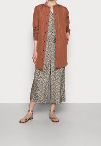 Quiltet i rustfarvet langt jakke med sorte trykknapper over en blomsterprint jumpsuit med brede ben, parret med minimalistiske bronze sandaler.