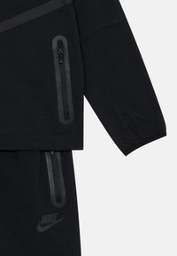Črni Nike trenirka z zadrgo na jakni in hlačah, z gladko teksturo in subtilnim logotipom v mat finišu.
