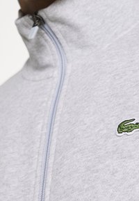 Ljusgrå zip-jacka i texturerad tyg, med stående krage och en grön broderad logotyp på vänster bröst.