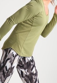 Langärmliges Sportoberteil in Olivgrün, aus texturiertem, atmungsaktivem Stoff. Kombiniert mit gemusterten Leggings in Schwarz, Grau und Beige.