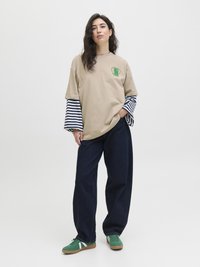Beige übergroßes T-Shirt mit grünem Grafikdruck, über einem gestreiften Langarmshirt getragen. Dunkelblaue weit geschnittene Jeans und grüne Sneakers mit weißen Streifen.