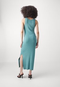Abito lungo teal a coste con spacco laterale, senza maniche e silhouette aderente. Il materiale sembra morbido con una texture liscia.