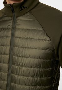 Lähetä upotettu vihreä takki lähellä, jossa on vetoketjukiinnitys ja päällystetty tekstiili yhdellä puolella, korostaen urheilullista ja modernia muotoilua.