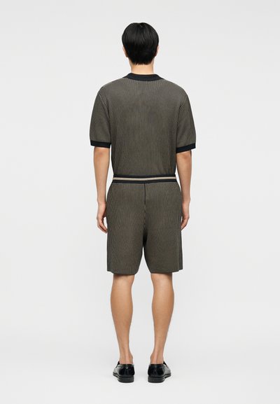 Άνδρας που στέκεται με την πλάτη προς τα εμπρός, φορώντας ένα σετ με ριγέ κοντομάνικο πουκάμισο και σορτς που ταιριάζουν, μαζί με μαύρα δερμάτινα παπούτσια.