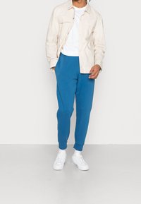 Ljusbeige skjortjacka över en vit t-shirt, ihopparad med blåa sweatpants och vita sneakers. Slät tyg och avslappnad passform.