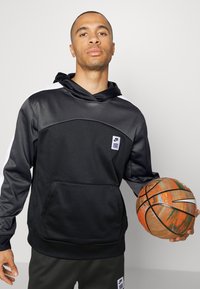 Felpa con cappuccio nera dalla consistenza liscia, con accenti bianchi sulle maniche e una tasca frontale. La persona tiene in mano un pallone da basket multicolore.
