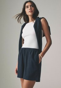 Top a maniche corte aderente bianco abbinato a pantaloni corti blu scuro con vita elasticizzata e tasche laterali; strato esterno drappeggiato.