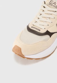 Sneaker krémbeige felsőrésszel, hálós textúrával és barna bőr részletekkel. Jellemzője a logó a sarkon, gumitalp és fűzős zárás.