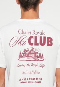 Hvit bomulls T-skjorte med en burgunder grafikk av en hytte, furu-trær, og teksten: "Chalet Royale Ski CLUB," med ytterligere kontaktopplysninger.