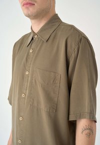 Kurzärmliges khakifarbenes Hemd aus weichem Stoff, mit einer durchgehenden Knopfleiste, spitzem Kragen und einer Brusttasche mit subtiler Textur.