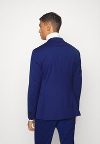 Marineblauer Anzug mit taillierter Passform, ausgestattet mit einem Revers und zwei Rückenschlitzen, kombiniert mit einem weißen Hemdkragen, der hinten sichtbar ist.