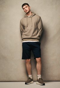 Hellbraun strukturierter Kapuzenpullover mit Kordelzügen, über einem weißen Shirt getragen; marineblaue Shorts mit Seitentaschen; braune Sneakers mit beigen Akzenten.