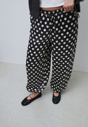 Personne portant un pantalon noir ample à pois blancs et des ballerines Mary Jane noires pailletées, debout sur un sol gris clair.