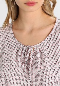 Weiße Bluse mit kleinem rotem Blumenmuster, rundem Ausschnitt und gebundenem Verschluss vorne. Leichter Stoff mit glatter Textur.