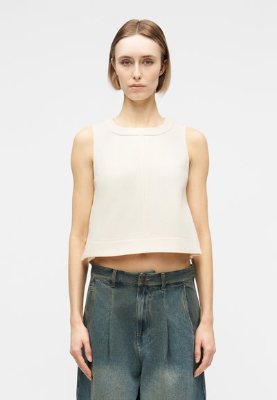 Femme portant un haut court blanc sans manches et un jean bleu ample taille haute, debout devant un fond clair uni.