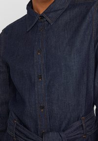 Mörkblå denimskjorta med krage, knappad framtid och ett tygbälte. Har sömnadsdetaljer och knappringar.