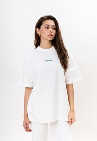 Witte oversized t-shirt van katoen, met in het midden op de borst de groene logotekst "MIRONS". Korte mouwen, eenvoudig, ontspannen pasvorm.