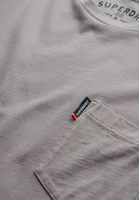 Grijze katoenen t-shirt met een ronde halslijn, voorzien van een borstzak en een klein zwart etiket met rode accenten. Zachte textuur, casual ontwerp.