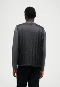 Μαύρο αμάνικο μπουφάν με γυαλιστερή υφή και ανοιχτή πλάτη, φορεμένο πάνω από μακρυμάνικη γκρι μπλούζα, με απλό, μοντέρνο σχέδιο.