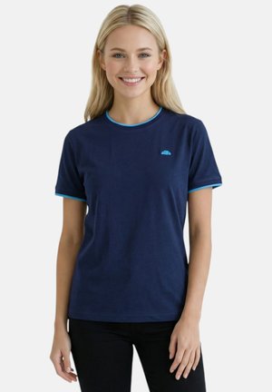 Femme souriante aux longs cheveux blonds, portant un t-shirt bleu marine avec bordure bleu clair et un pantalon noir, debout sur un fond blanc.