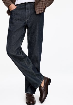 Personne portant un jean en denim foncé, des mocassins en cuir marron, une chemise blanche et une veste marron à carreaux, les mains dans les poches.