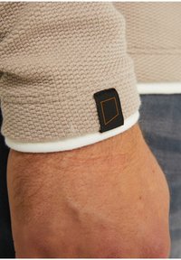 Bežová texturovaná pletenina s bílým manžetou. Obsahuje černou nášivku s oranžovým okrajem blízko zápěstí, což zdůrazňuje detaily oděvu.